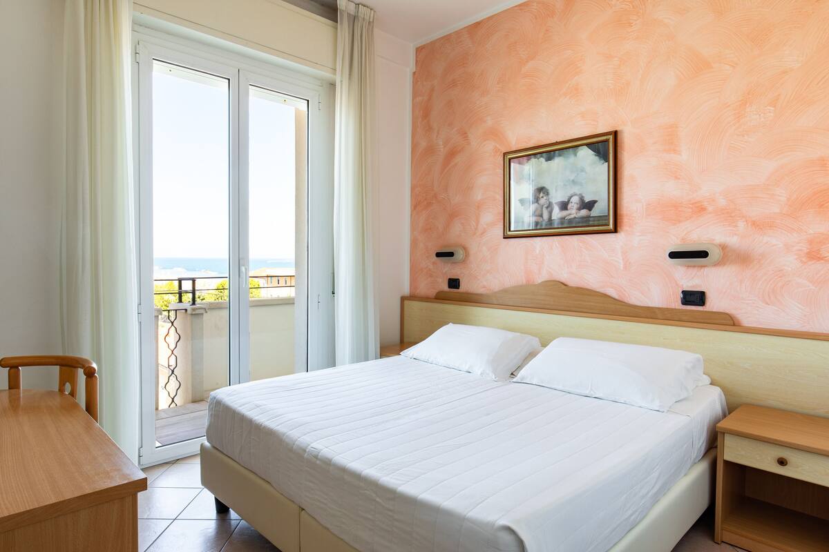 Camera d'albergo con un letto, un comodino in legno, una scrivania e vista sul mare attraverso una porta scorrevole in vetro.
