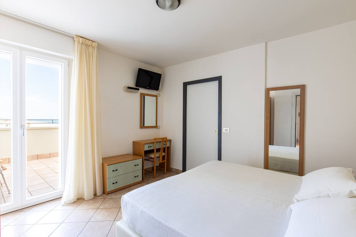 Camera d'albergo con un letto, una scrivania, una sedia, una TV, uno specchio e una porta-finestra scorrevole su un balcone.
