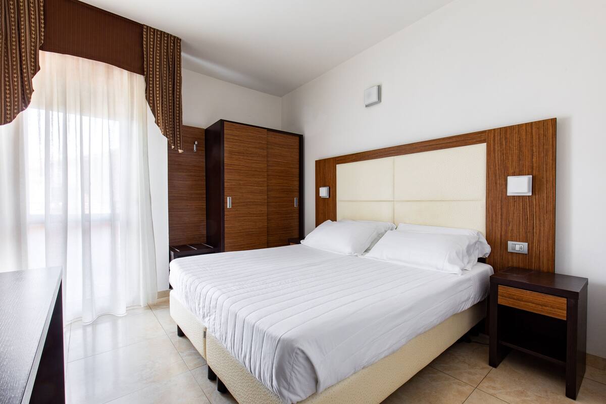 Una camera da letto ordinata con un letto grande, un armadio in legno, un comodino e una finestra con tende trasparenti.