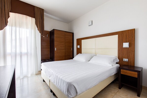 Un dormitorio ordenado con una cama grande, un armario de madera, una mesita de noche y una ventana con cortinas traslúcidas.