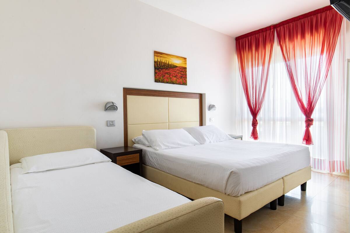 Una camera d'albergo con due letti, una testiera, un comodino, un quadro e tende rosse.