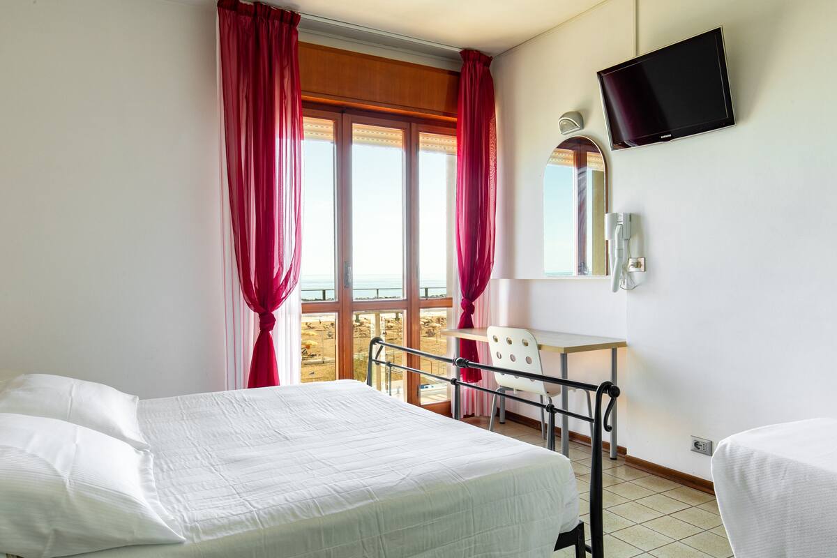 Una camera d'albergo con un letto, una scrivania, una sedia, una televisione e una finestra con tende.
