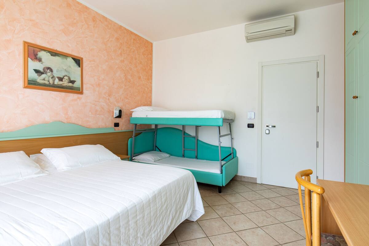 Una stanza con un letto a castello, un letto singolo, un tavolo in legno, una porta e un condizionatore d'aria a parete.