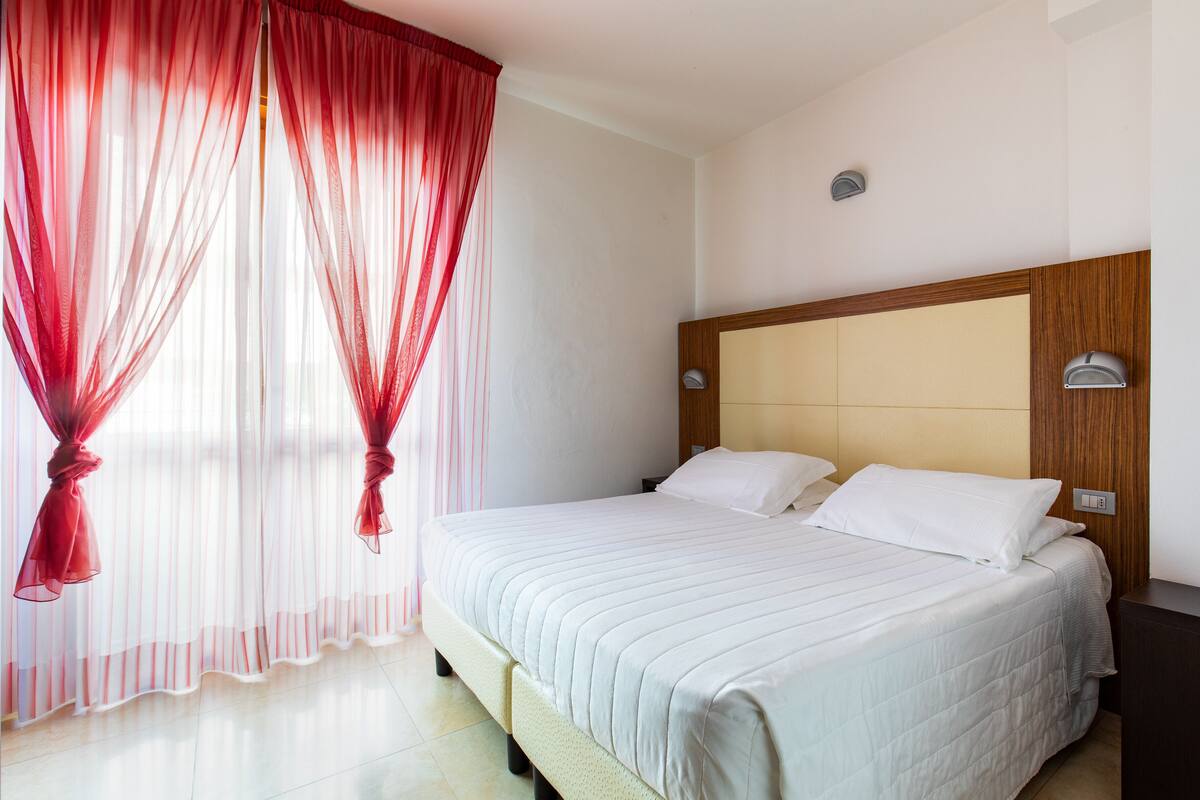 Camera d'albergo con un letto grande, una testiera in legno e tende rosate trasparenti.
