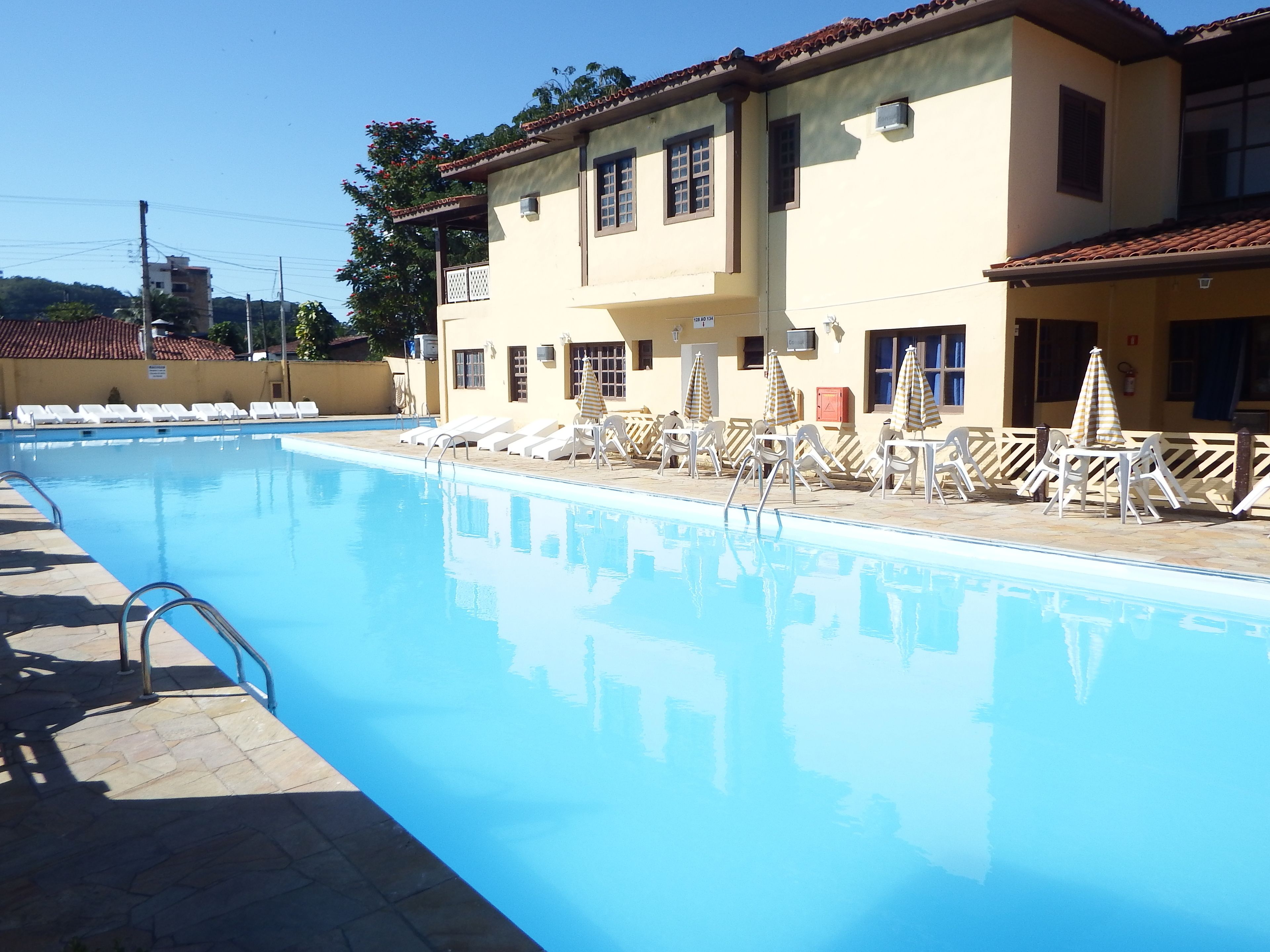 Piscina com espreguiçadeiras e guarda-sóis, rodeada por um edifício com várias janelas e telhado em azulejos.