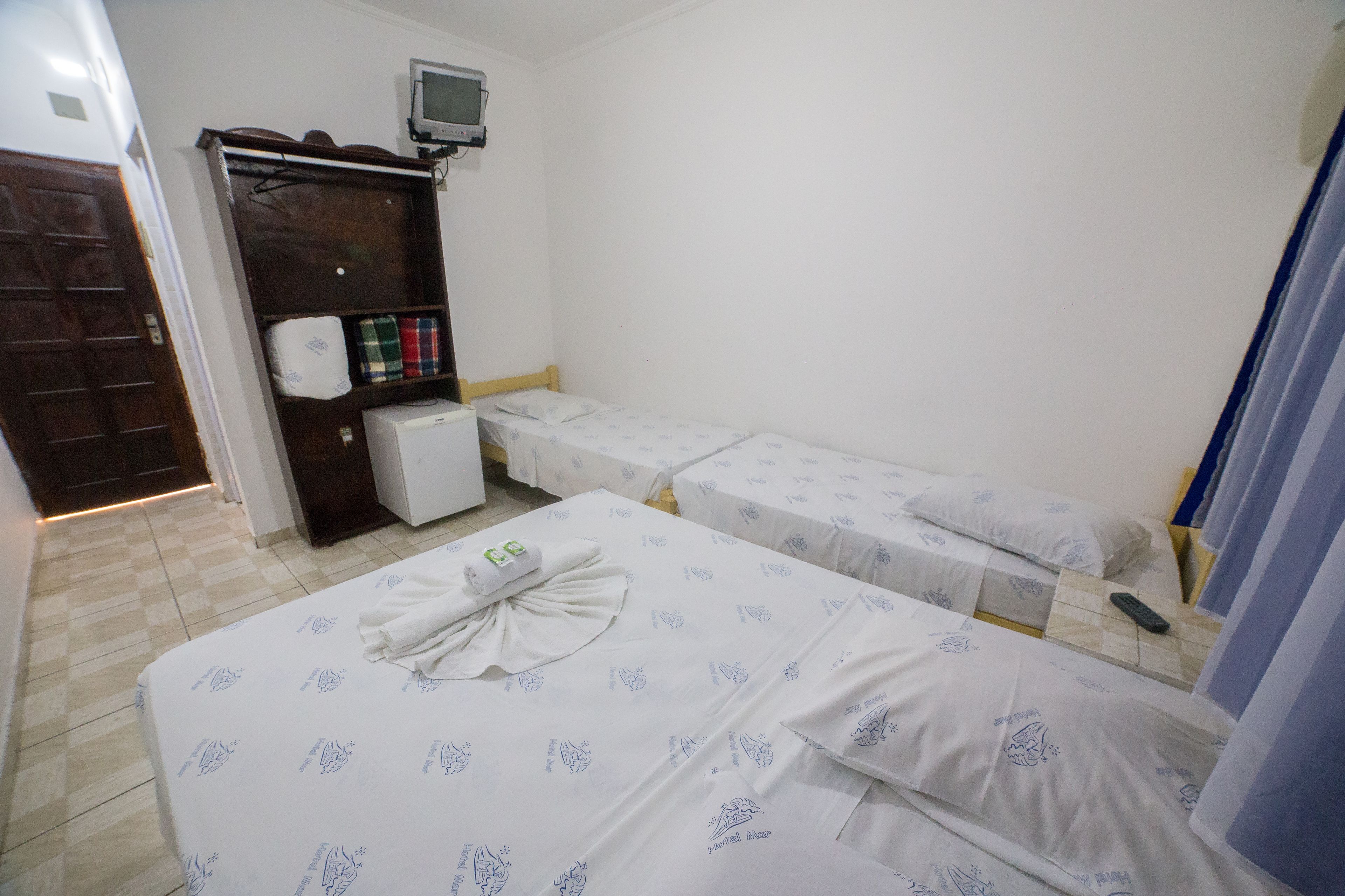 Quarto de hotel com duas camas de solteiro, uma pequena mesa com computador, um guarda-roupa e uma televisão na parede.