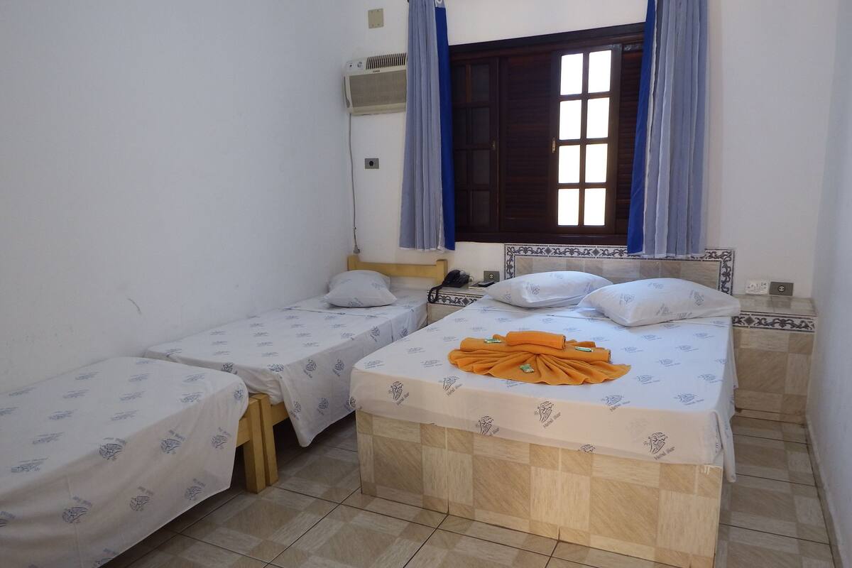 A small, clean room with two single beds, a wooden headboard, a small bedside table with a lamp, and a window with curtains.