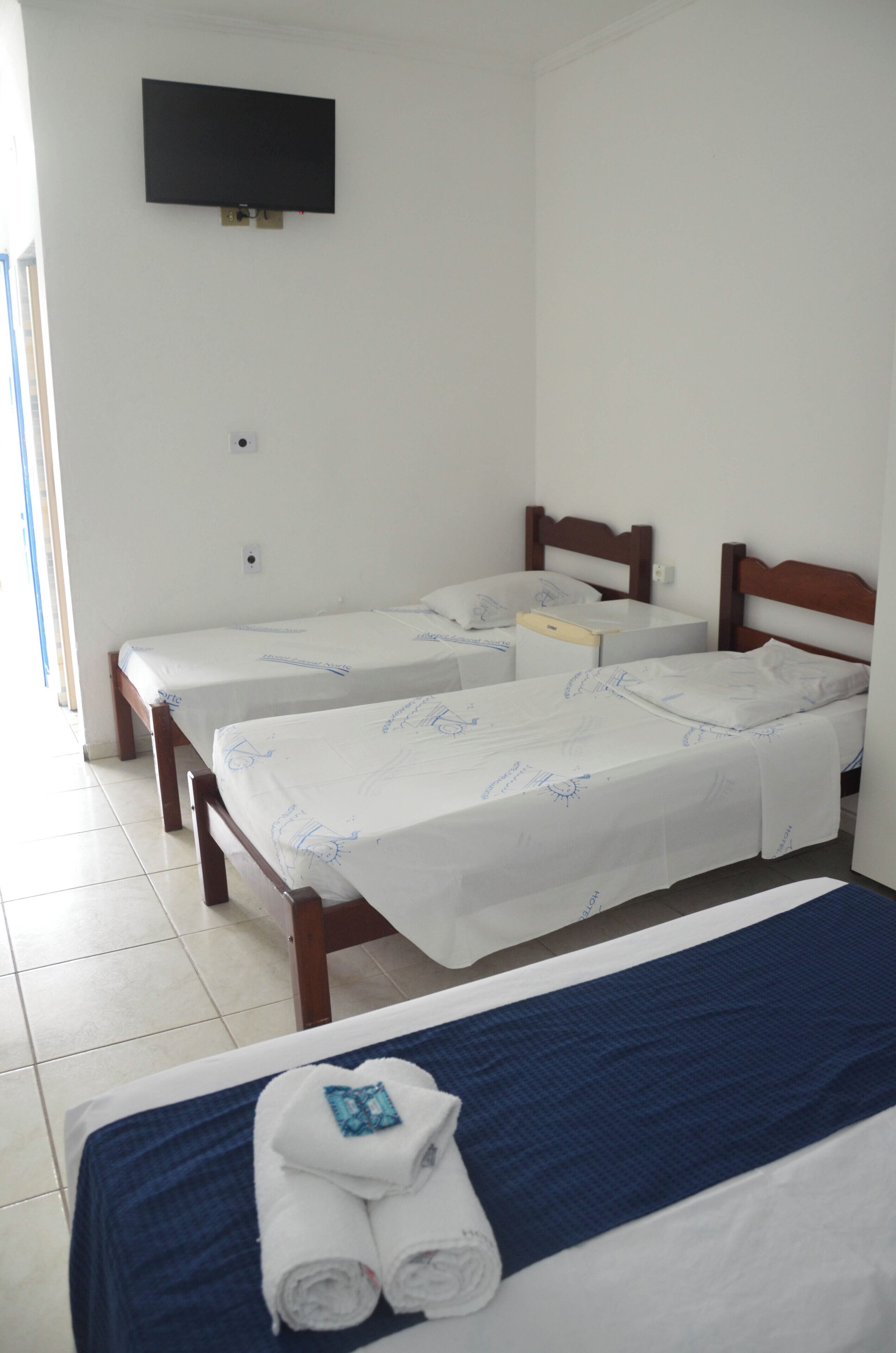 A hotel room with two single beds, a larger bed, a TV mounted on the wall, and a window providing natural light.