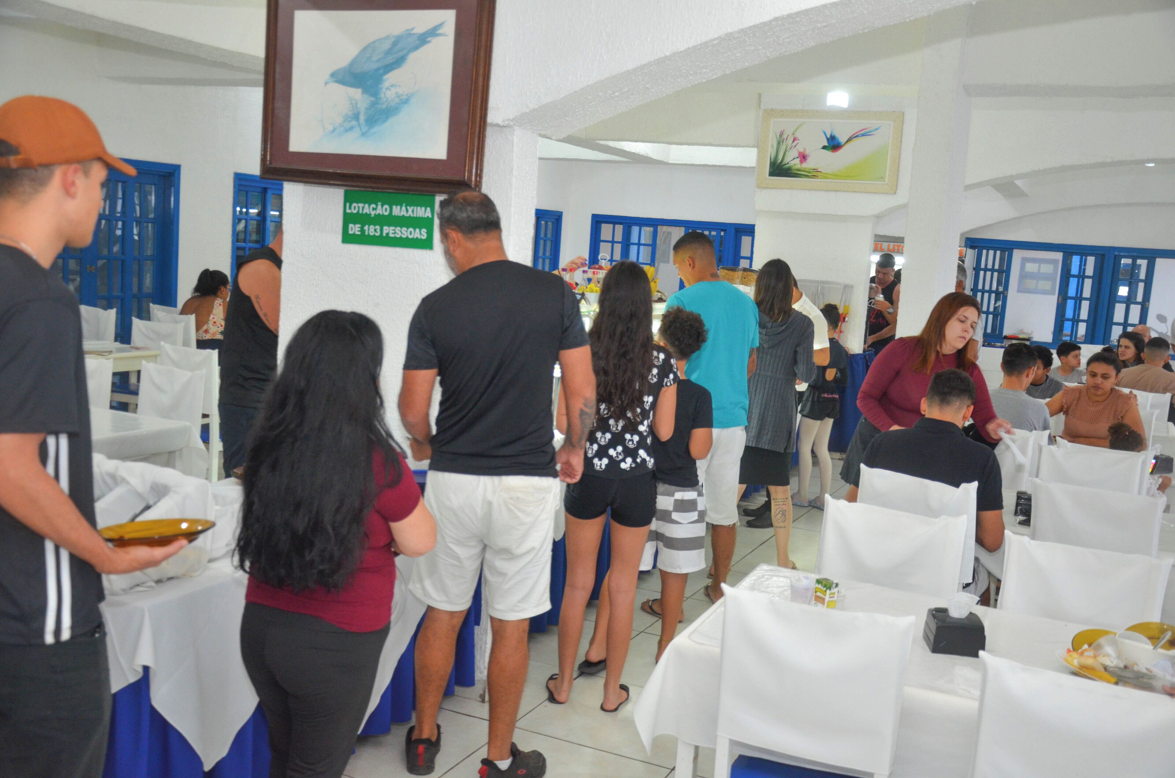 A crowded indoor event with tables and chairs arranged in rows, a sign indicating maximum capacity, and people standing and sitting.