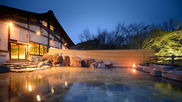 A traditional Japanese onsen with a stone bath surrounded by rocks, a wooden building with large windows, and a night sky.