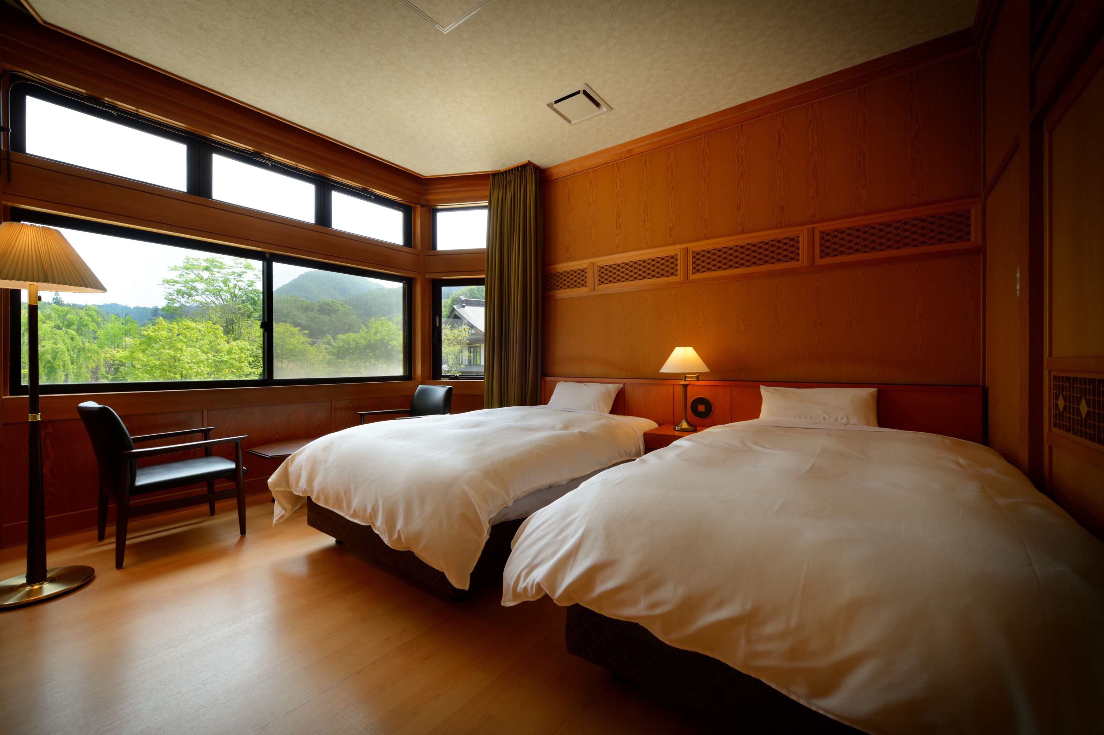 A hotel room with two beds, a chair, a lamp, and large windows offering a view of greenery.