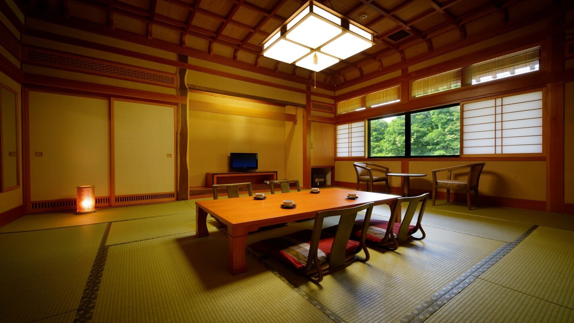 A traditional Japanese room with tatami flooring, a low table, chairs, a television, and a window with a view of trees.