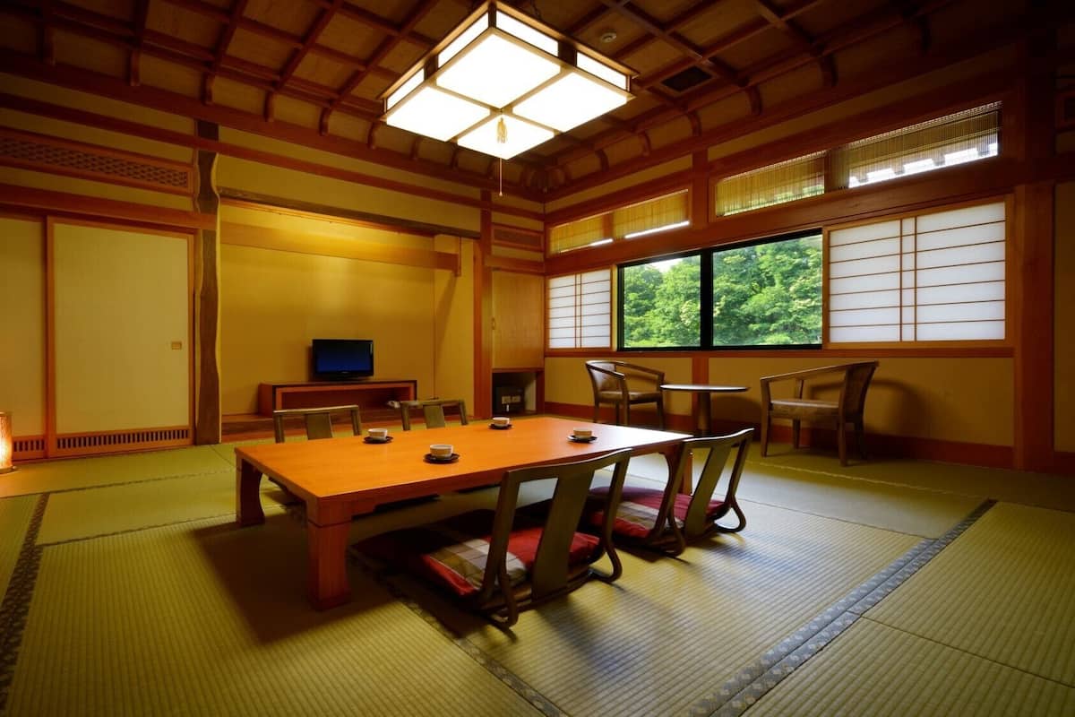 A traditional Japanese room with tatami flooring, a low table, chairs, a television, and a window with a view of trees.