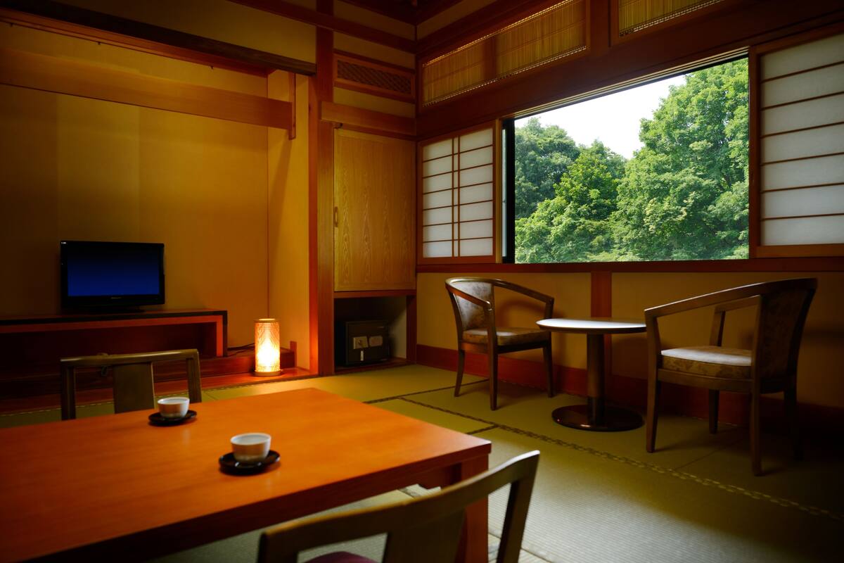 Ett traditionellt japanskt rum med tatamimattor på golvet, ett lågt bord med två kuddar, en tv och ett fönster med utsikt över en skog.