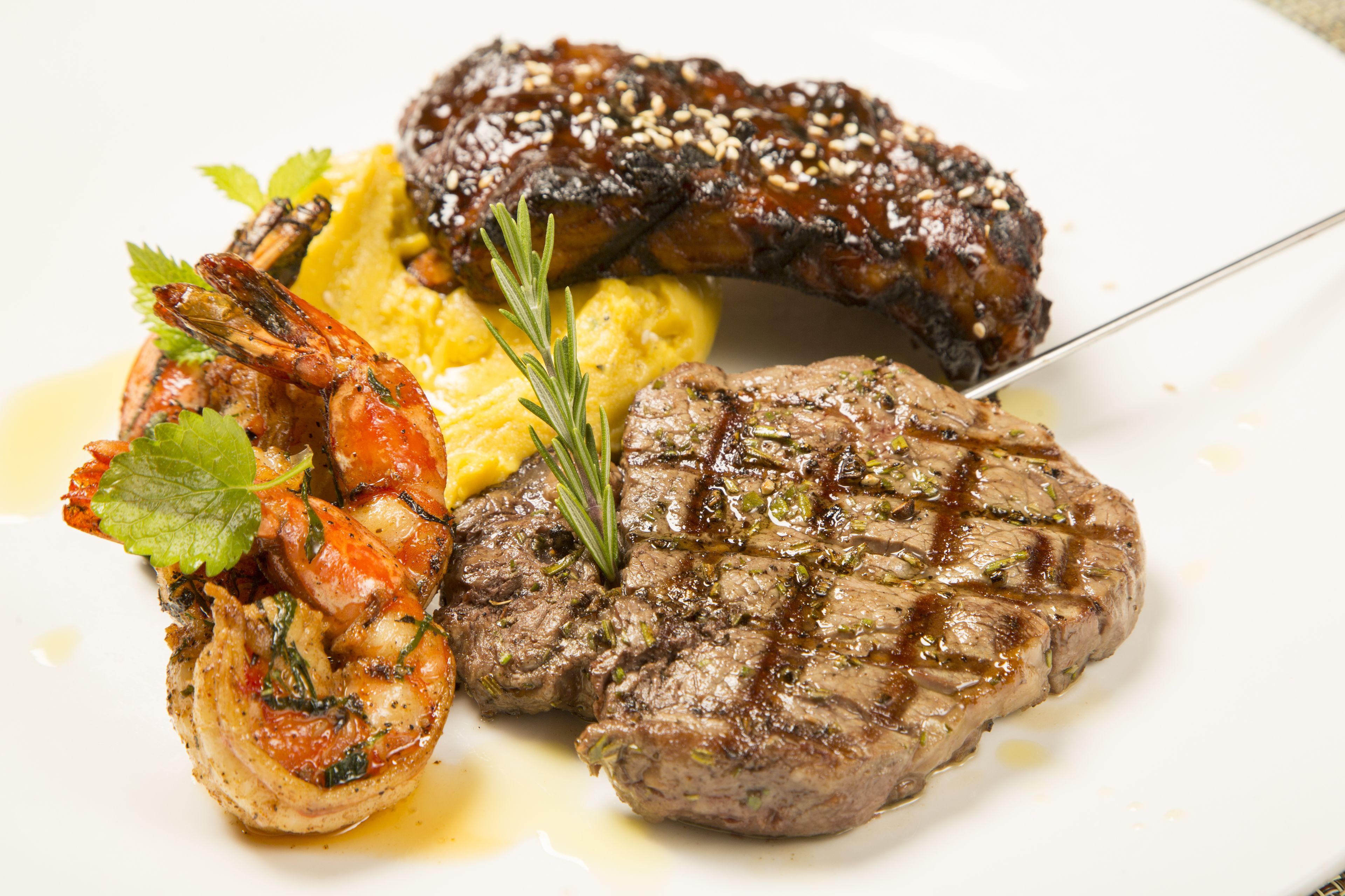 A plate with grilled steak, shrimp, and a sausage, garnished with herbs.
