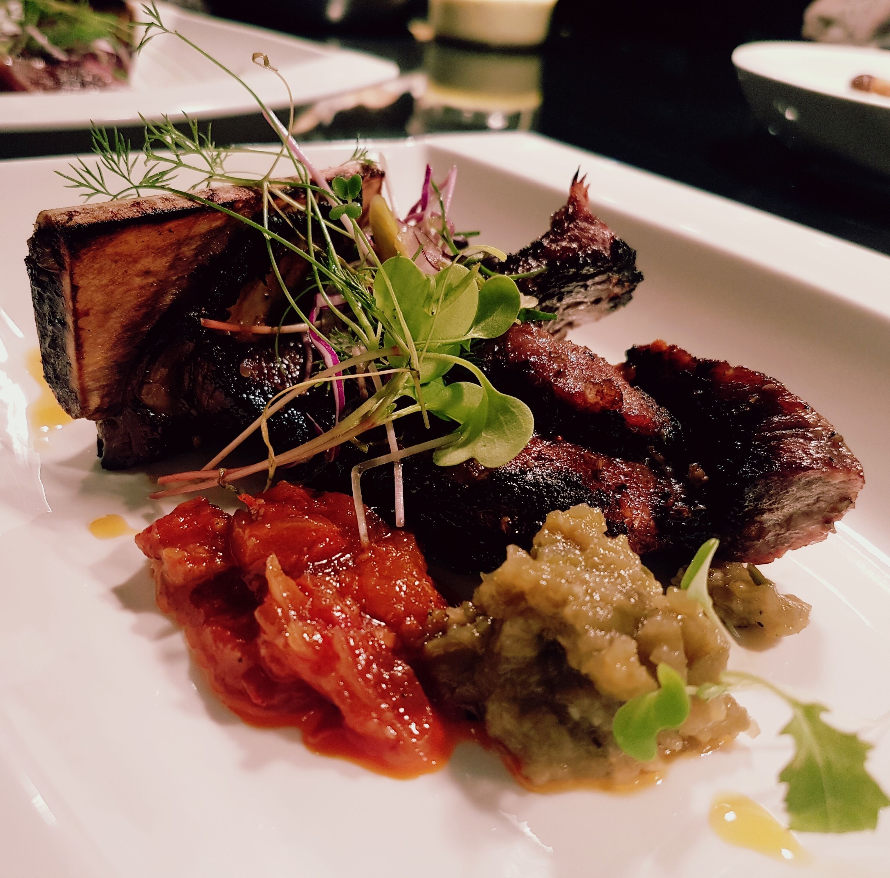 A plate of food with various dishes including a piece of meat, a red sauce, and a green garnish.