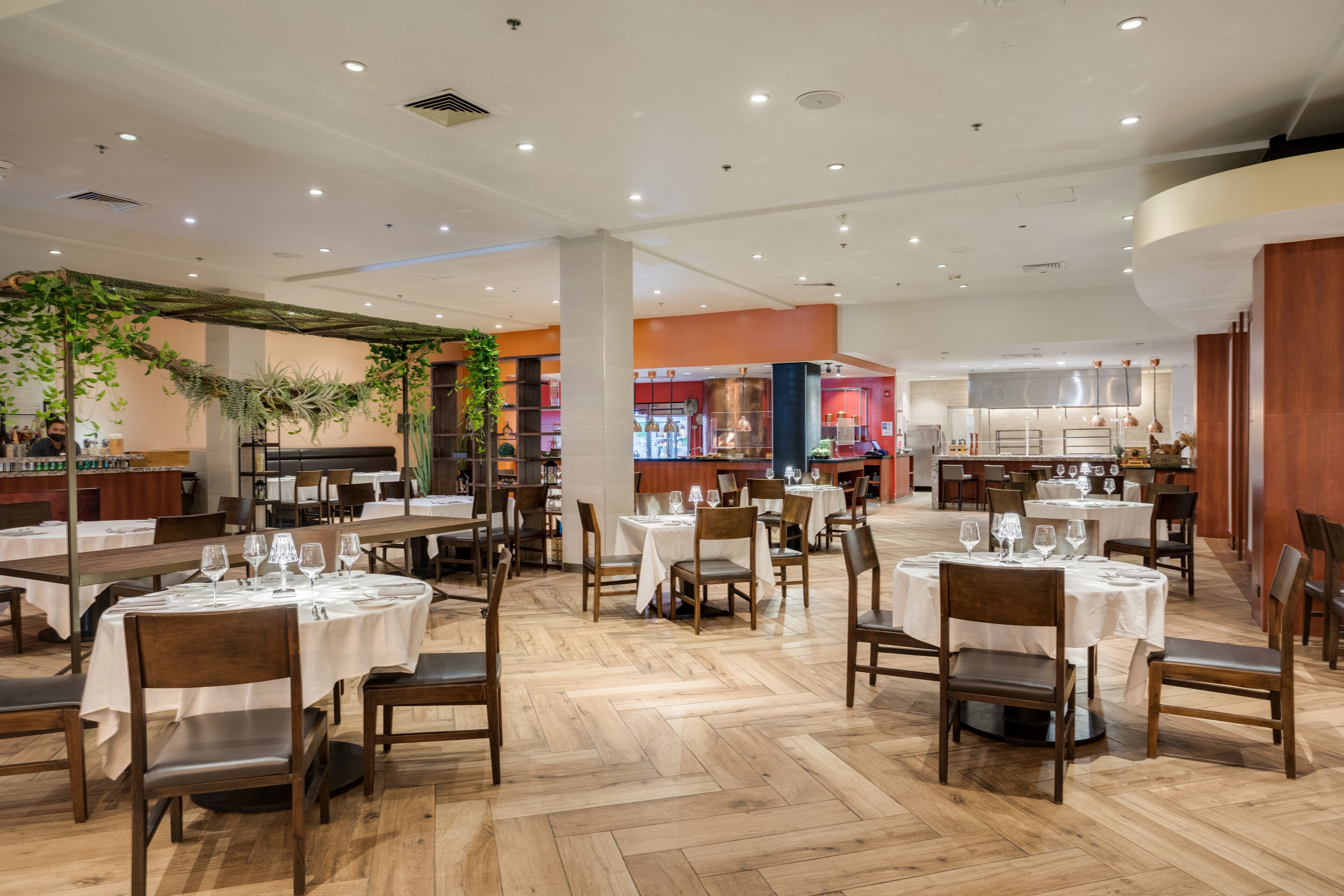 A spacious restaurant with multiple tables and chairs, a herringbone floor pattern, and a bar area in the background.