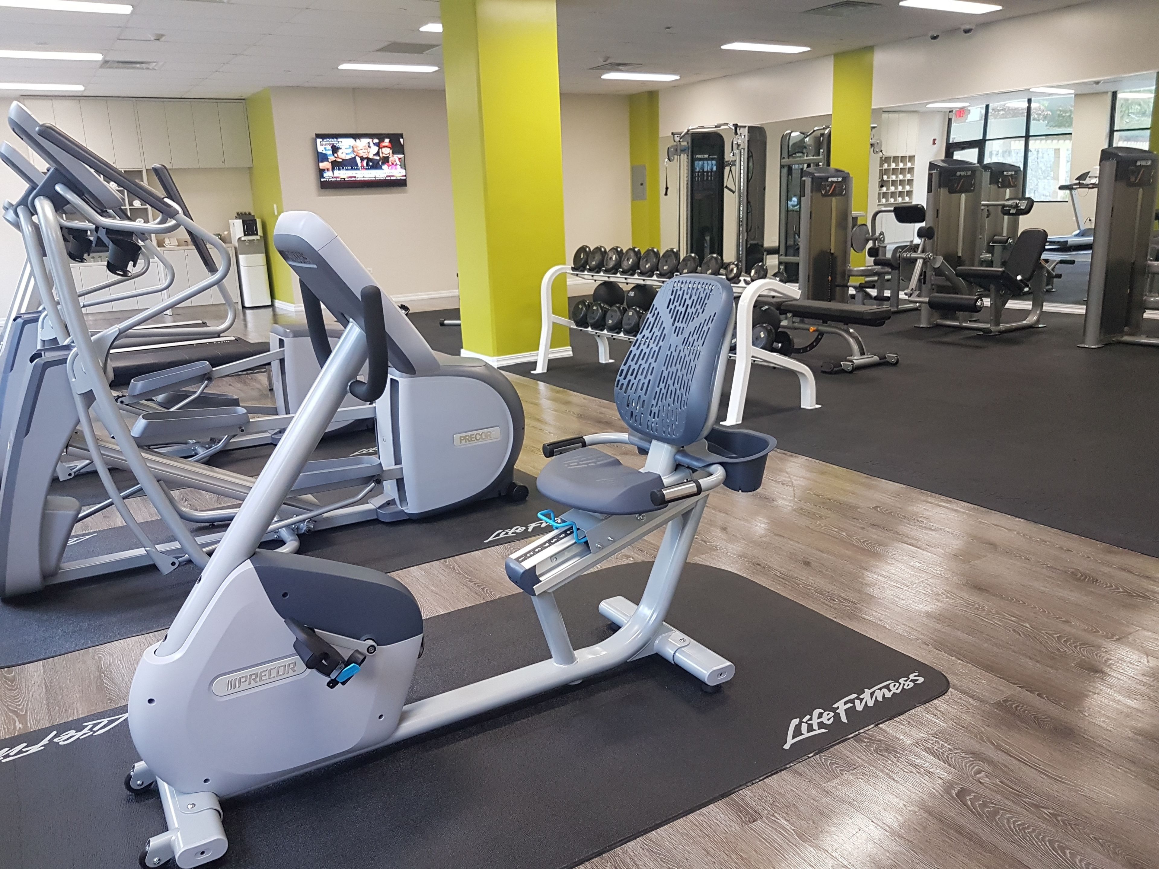 Academia com aparelhos de cardio, equipamentos de musculação e uma televisão na parede.