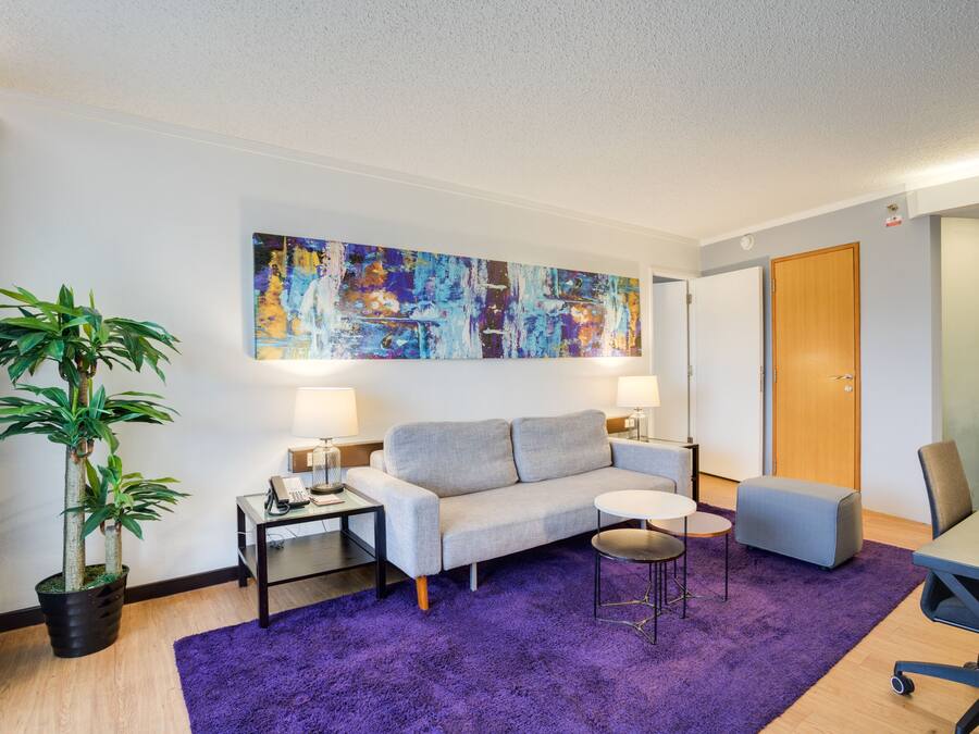 A modern living room with a grey sofa, a small table, a purple rug, and a large abstract painting on the wall.