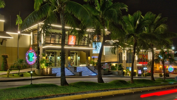 A modern hotel with a well-lit entrance, palm trees, and a staircase leading to the lobby. - Guam Plaza Resort (Tamuning)