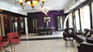 A spacious living room with a dining area, featuring a chandelier, ceiling fans, and large windows. - Pulse Impulse (New Delhi)