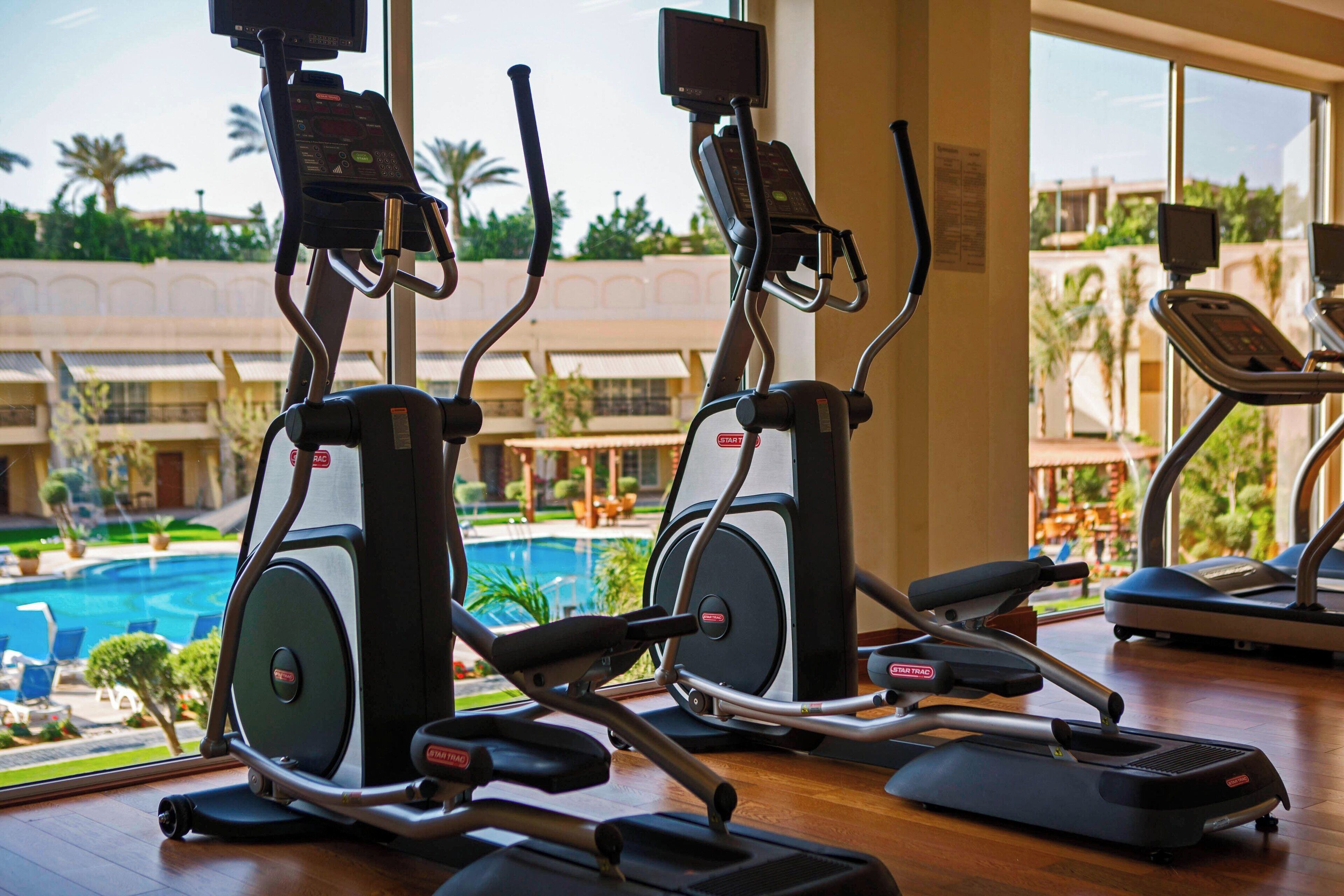Un gimnasio con cintas de correr y equipo de ejercicio, con vistas a una piscina y palmeras.