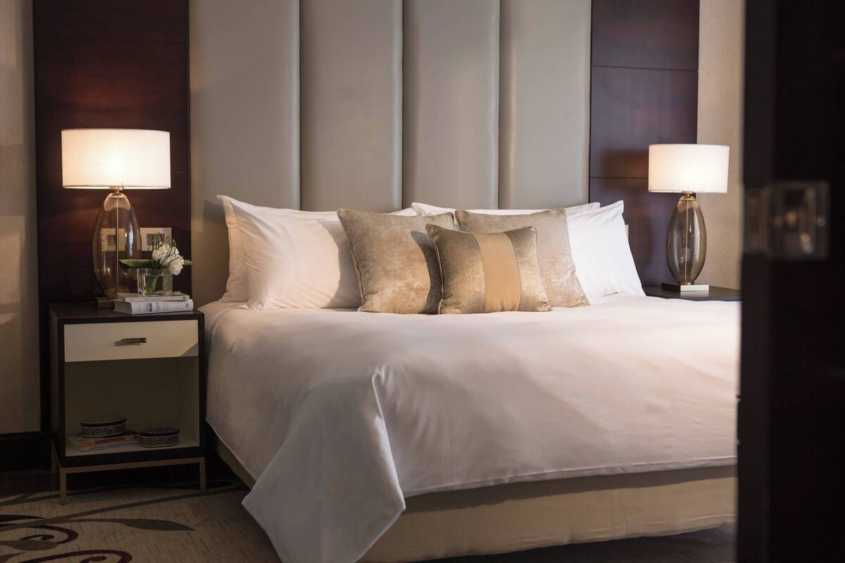 A neatly made bed with white linens and multiple pillows, flanked by two bedside tables with lamps, in a modern hotel room.