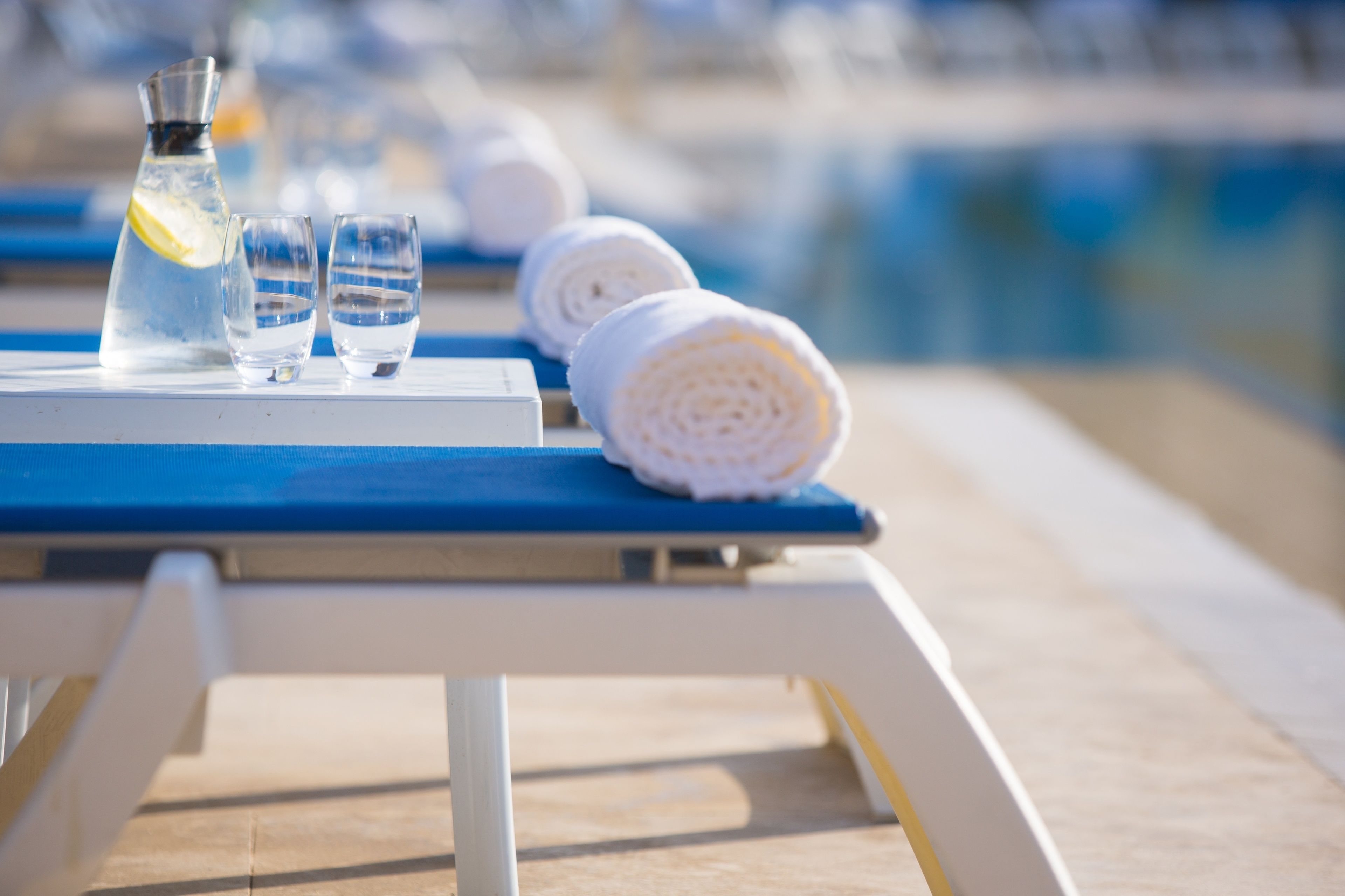 A poolside setting with a table holding a water bottle, two glasses, and rolled towels.