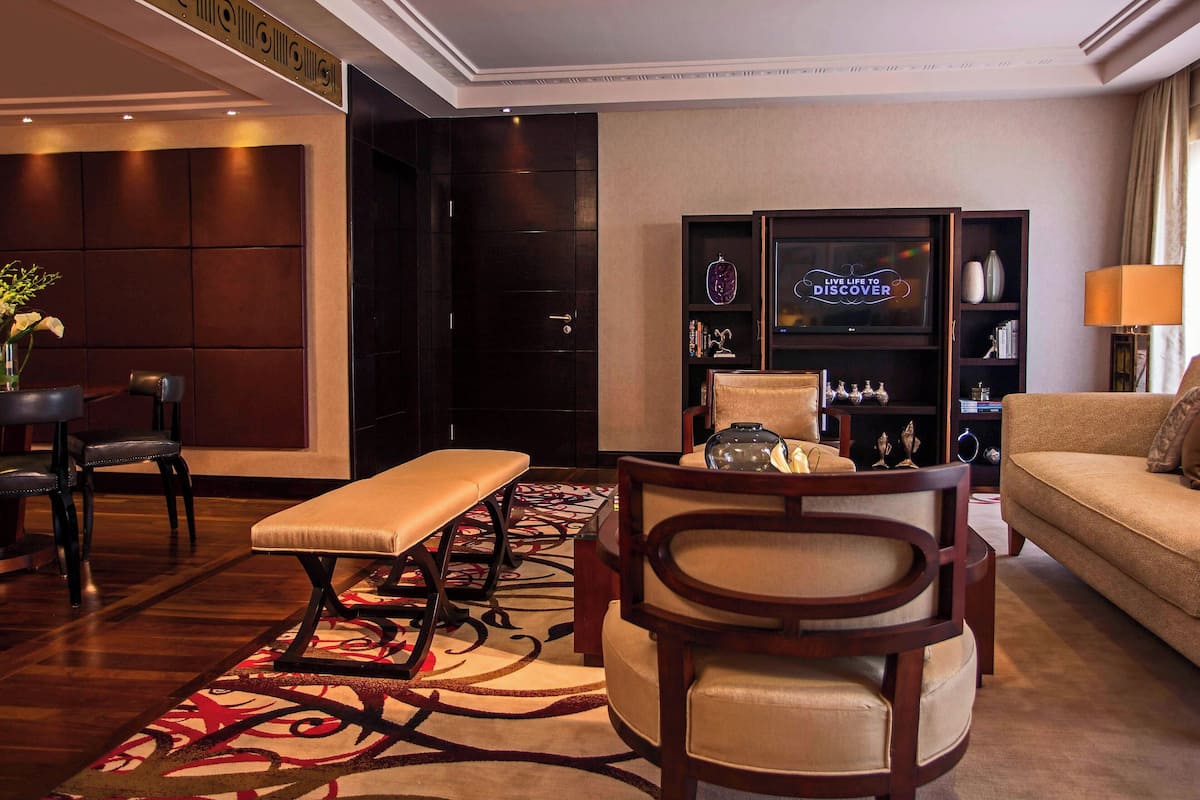 A modern hotel lobby with a sofa, a bench, a dining table, and a television displaying "LIVE LIFE TO DISCOVER".