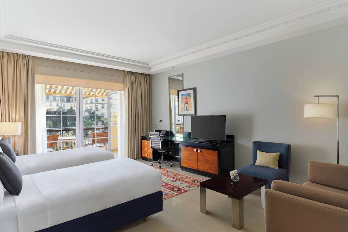 A hotel room with a bed, a desk, a chair, a TV, and a balcony with a view of buildings.