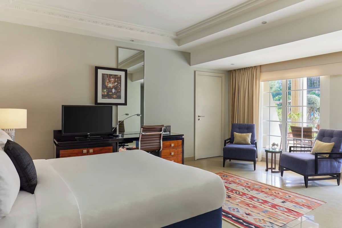 A hotel room with a large bed, a desk with a television, two armchairs, and a large window with curtains.