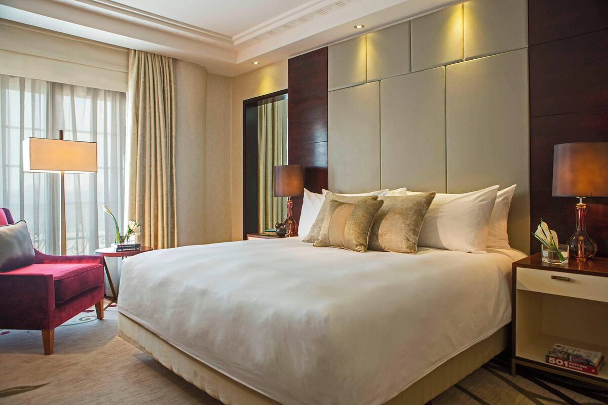 A large bed with multiple pillows, a bedside table with a lamp, a chair, and a nightstand with a lamp.
