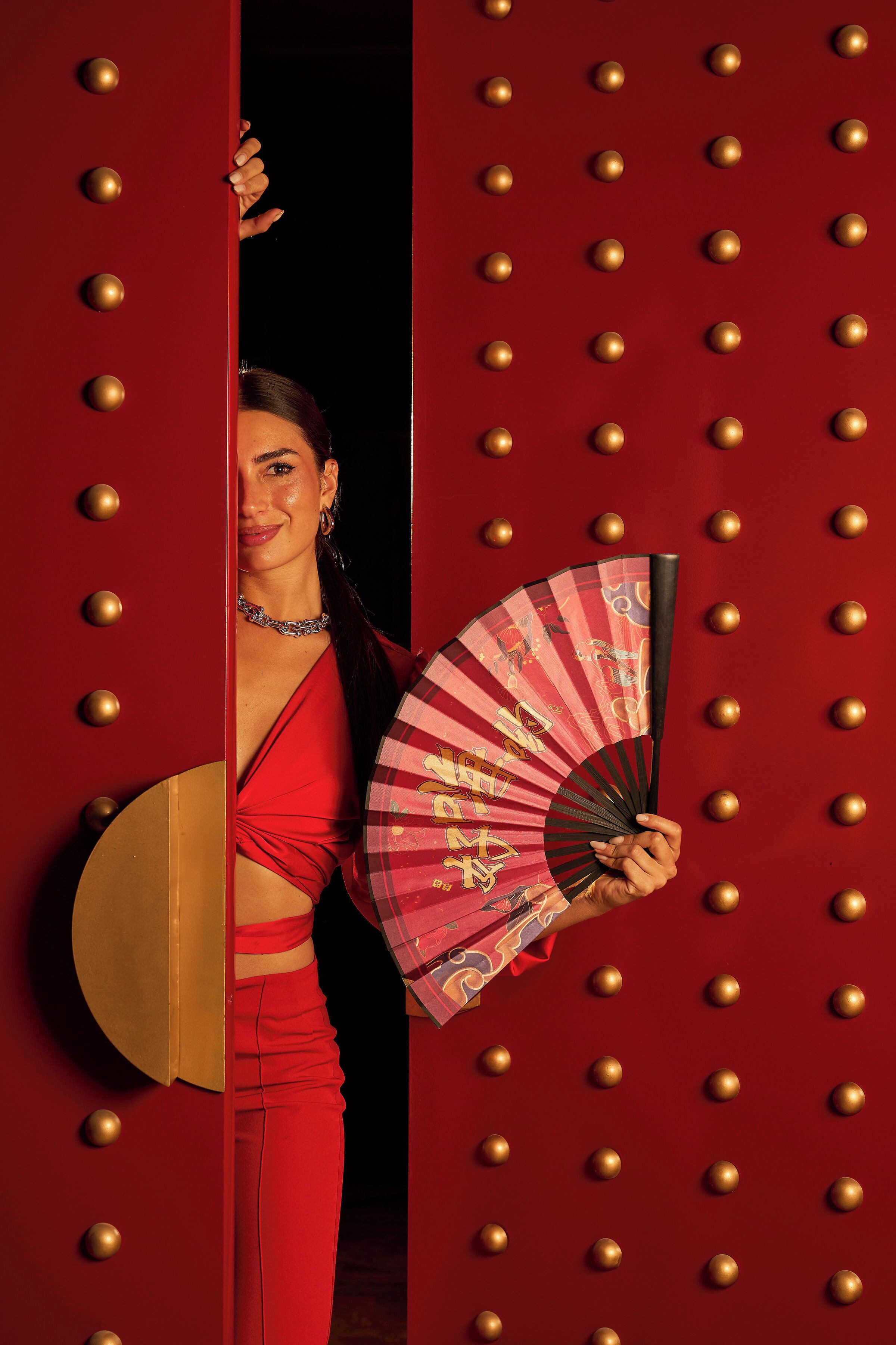 Una persona vestida de rojo sostiene un abanico y un objeto redondo, de pie frente a una puerta roja adornada con tachuelas doradas.