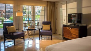 Une chambre d’hôtel comprenant un lit, deux fauteuils, une petite table avec une lampe, un téléviseur et donnant sur une piscine extérieure.