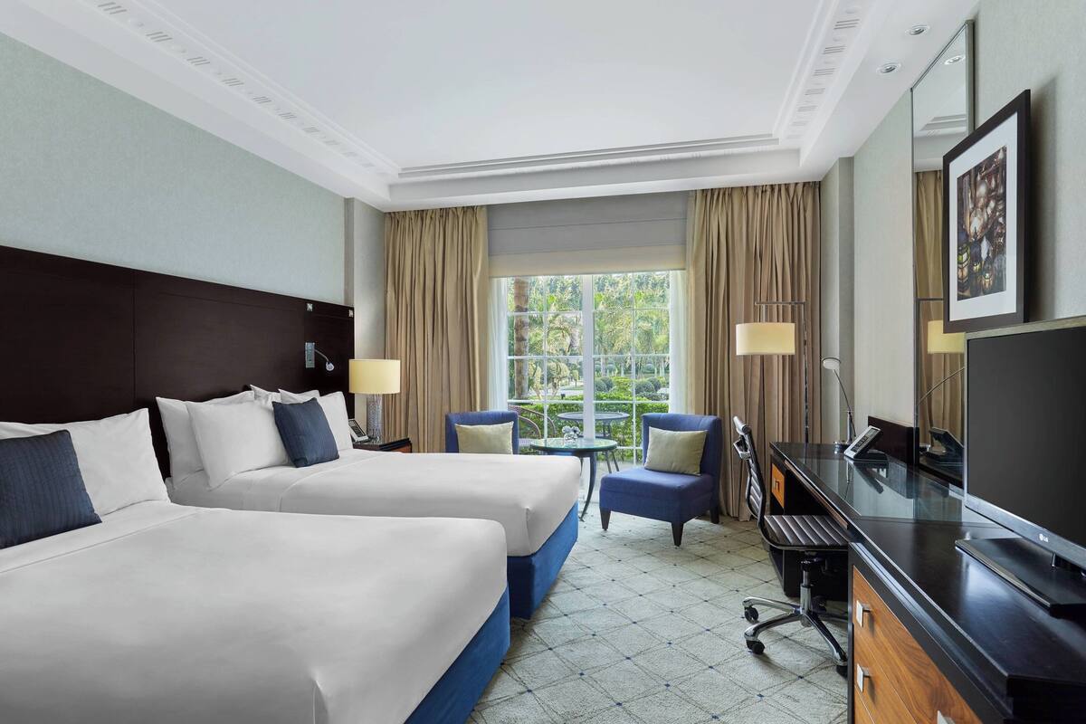 A hotel room with two beds, a desk with a computer, a TV, and a window with curtains.