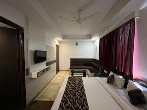 A hotel room with a bed, a television mounted on the wall, a ceiling fan, and a window with curtains. - Hotel Taj Galaxy  (Agra)
