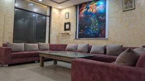 A room with a large sofa set and a coffee table, a large window, and a colorful peacock painting on the wall. - Hotel Taj Galaxy  (Agra)