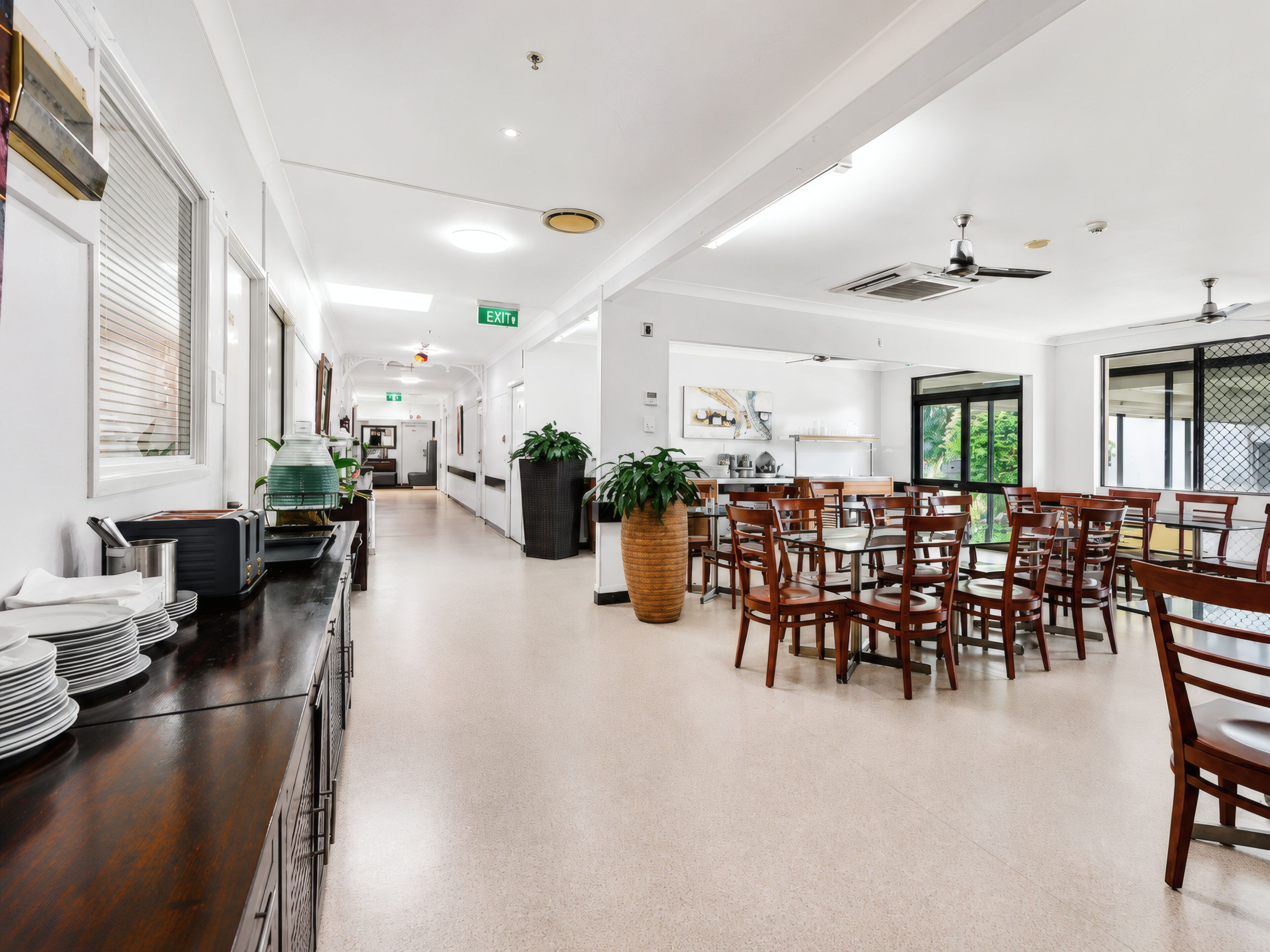 A spacious hotel dining area with a long counter, multiple tables, and chairs, a large potted plant, and a view through a window.