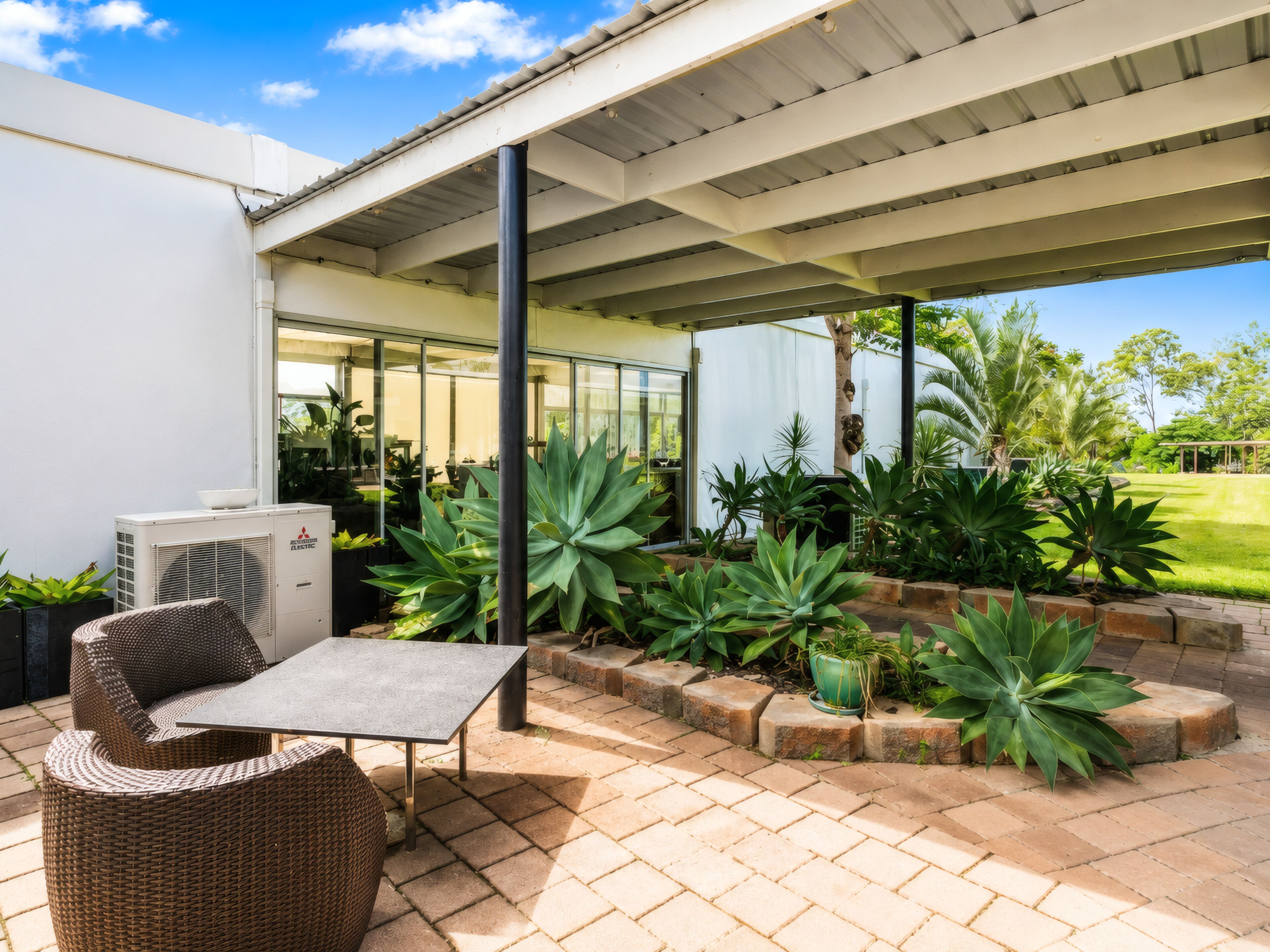 An outdoor patio area with a covered seating area, planters with large-leafed plants, and a modern building with large windows.