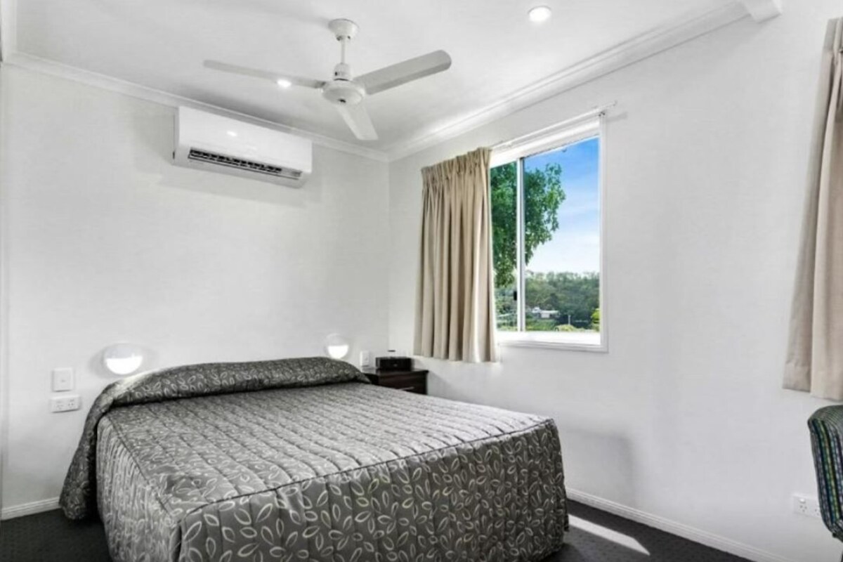Ein Hotelzimmer mit Bett, Klimaanlage, Deckenventilator, einem Fenster mit Vorhängen und Blick auf Grünfläche.