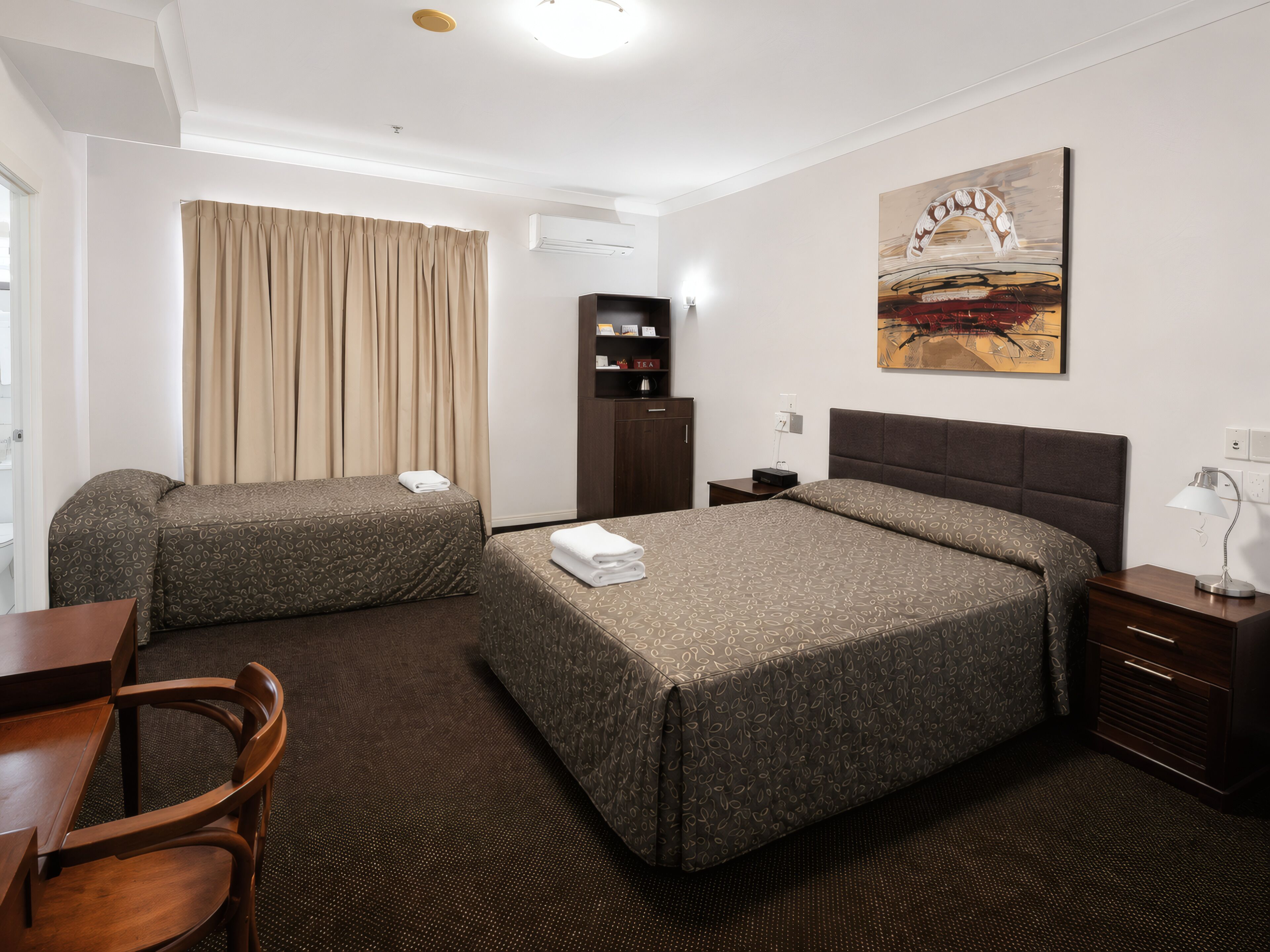 A hotel room with two beds, a desk, a chair, a nightstand, a lamp, a bookshelf, and a painting on the wall.