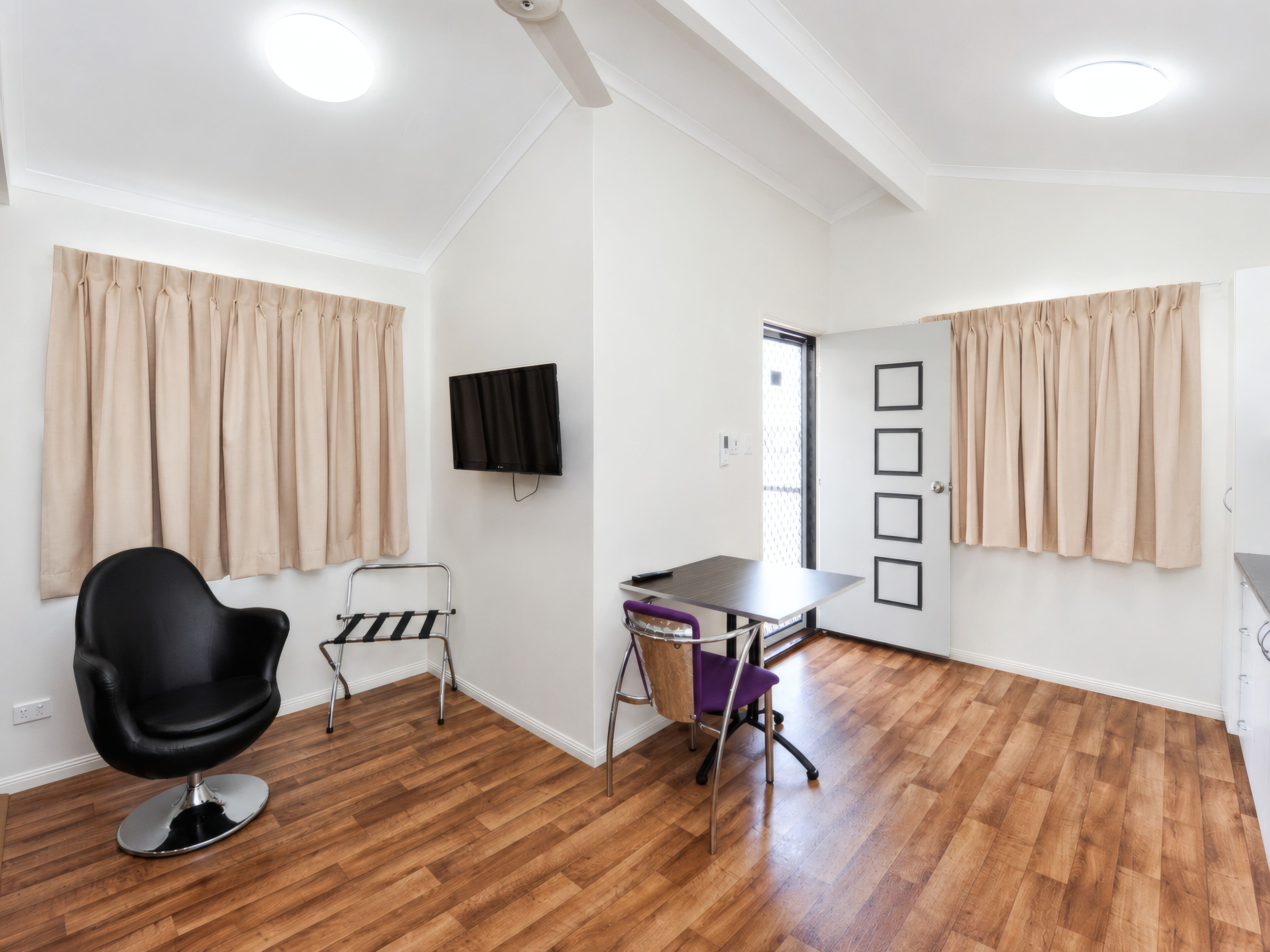 Una habitación con piso de madera, una silla negra, una mesita pequeña, un televisor instalado en la pared y cortinas color crema.