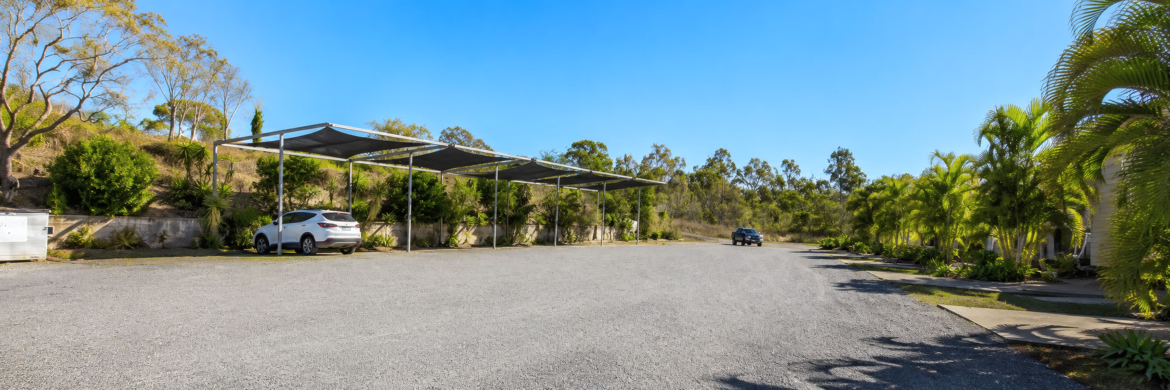 En parkeringsplads med et overdækket carport, en hvid bil og en bygning i baggrunden.