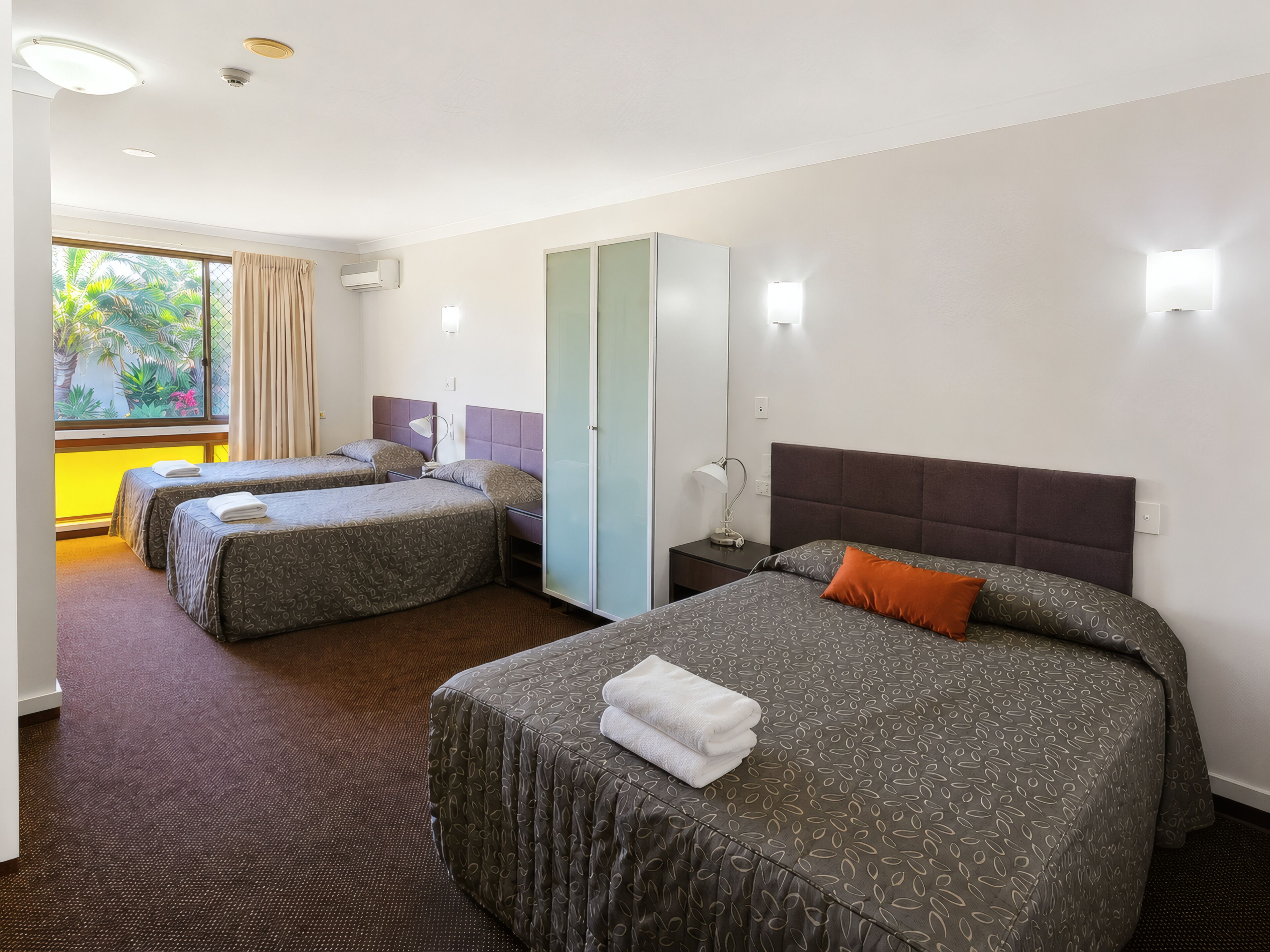 Ein Hotelzimmer mit zwei Betten, einem Kleiderschrank, einem Nachttisch und einem Fenster mit Vorhängen.