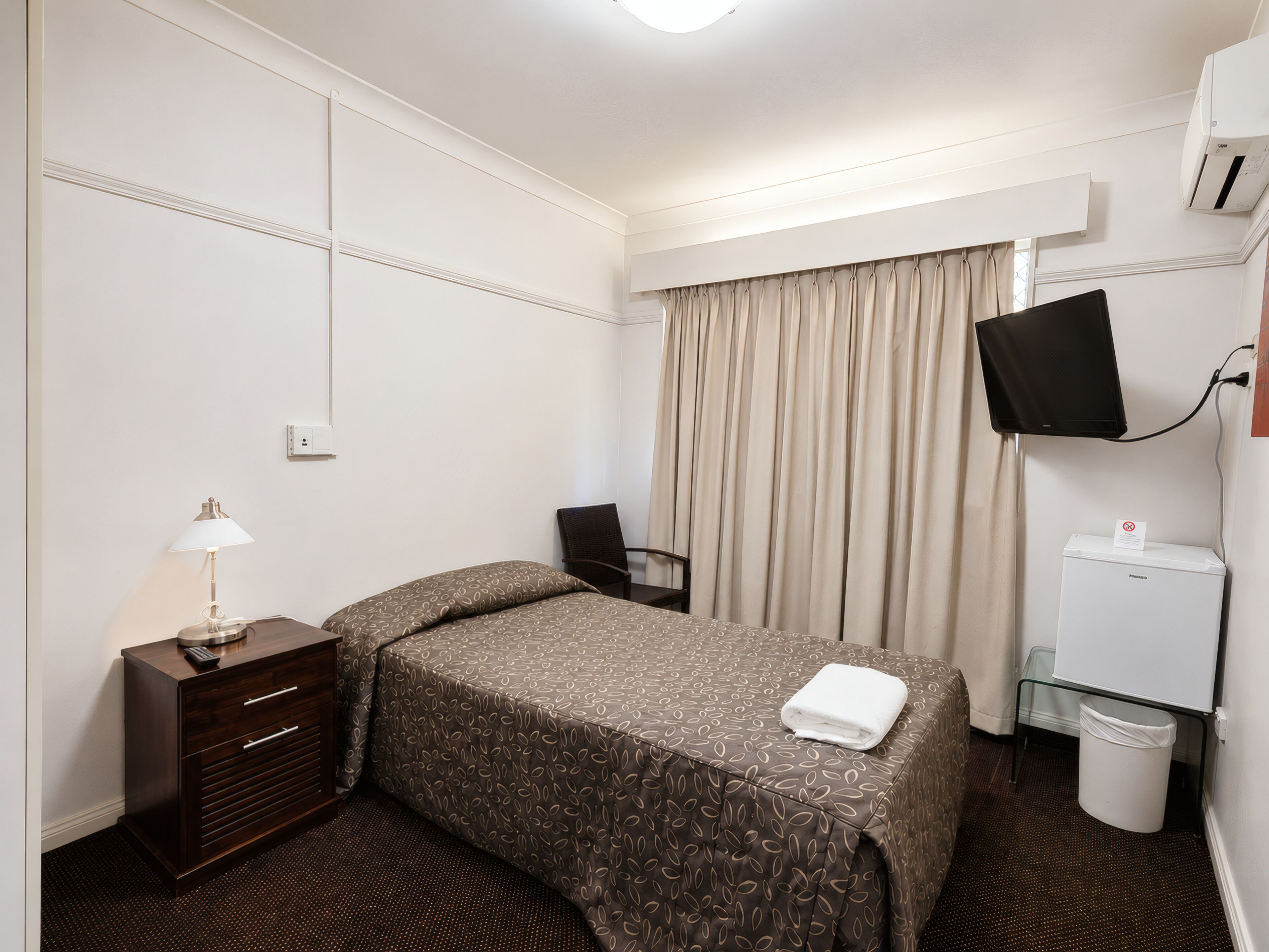 A hotel room with a bed, a desk, a chair, a TV, an air conditioner, and a trash bin.