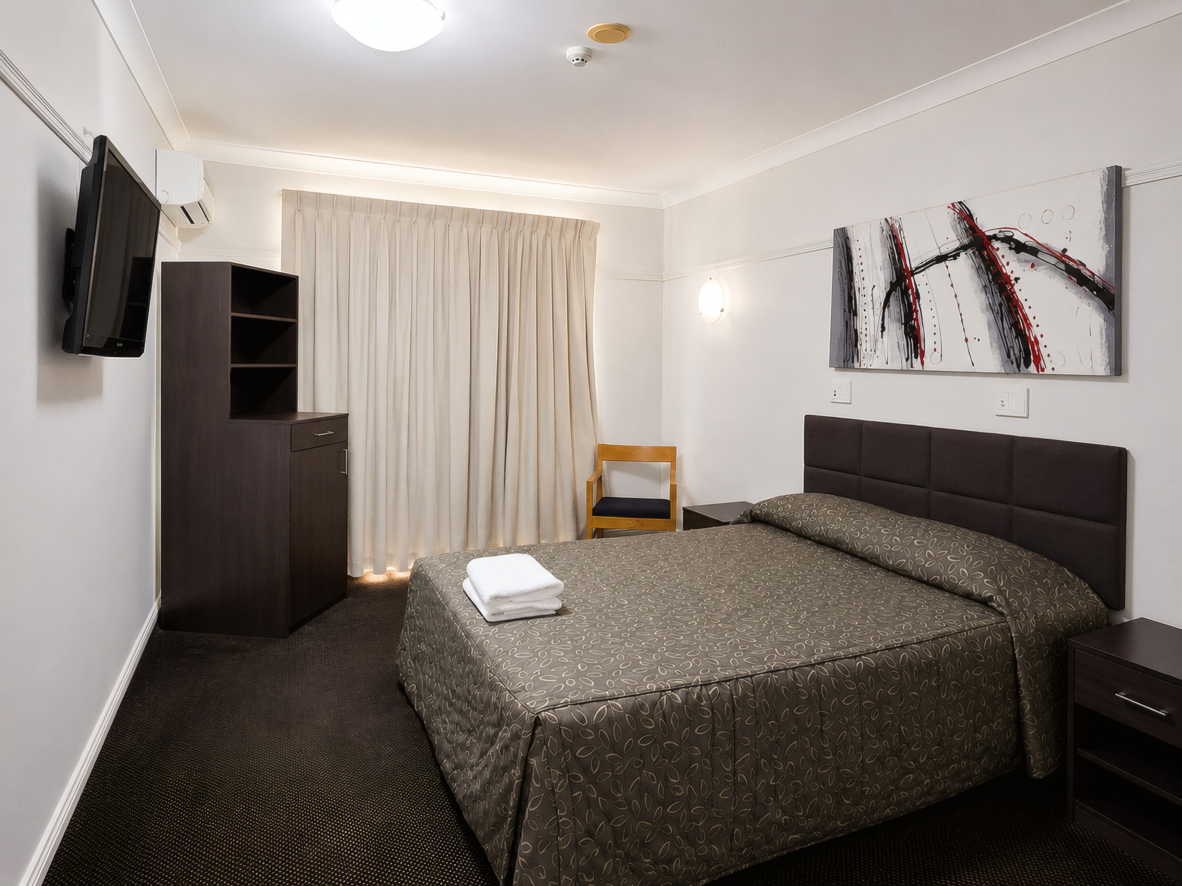 Une chambre d’hôtel avec un lit, une chaise, une armoire, un téléviseur à écran plat et un tableau au mur.
