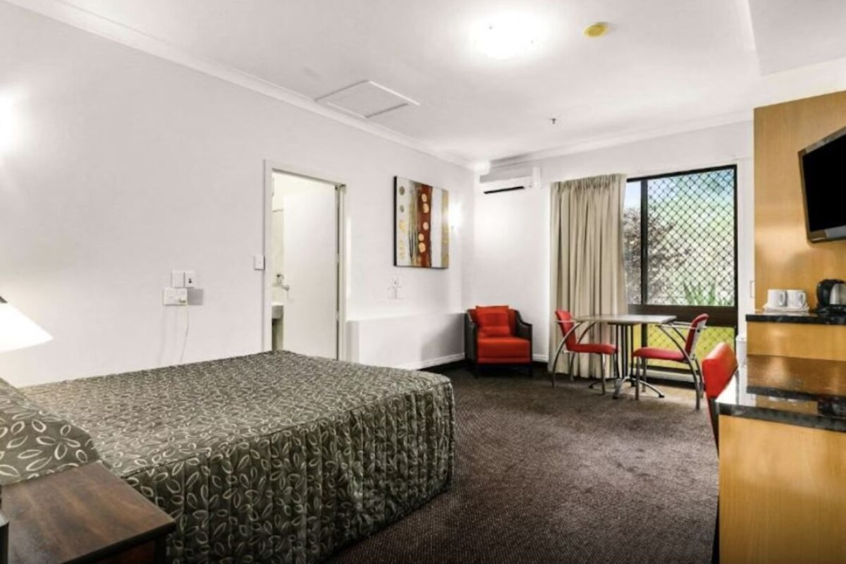 Une chambre d’hôtel comprenant un lit, un bureau avec une chaise, une petite table avec des chaises et une fenêtre avec des rideaux.