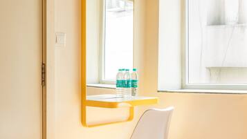 Une chambre d’hôtel avec un miroir à cadre jaune, une chaise blanche, une petite table avec des bouteilles d’eau et une fenêtre donnant sur des immeubles.