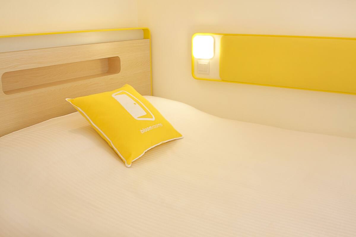 Een bed met een houten hoofdbord, een geel kussen en een nachtkastje met een lamp.