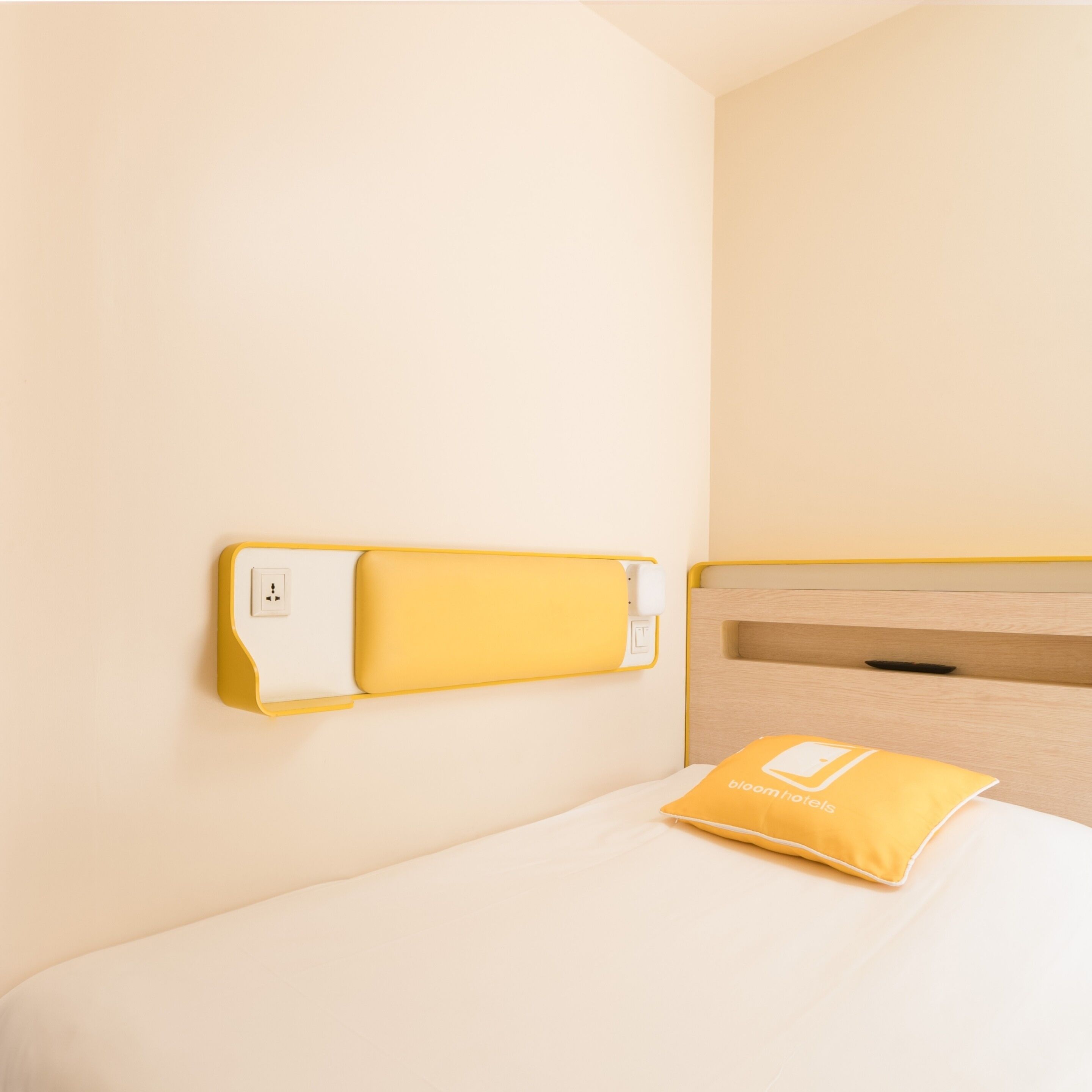 Hotelkamer met een bed, nachtkastje, een wandcontactdoos en een geel kussen met het logo van het hotel.